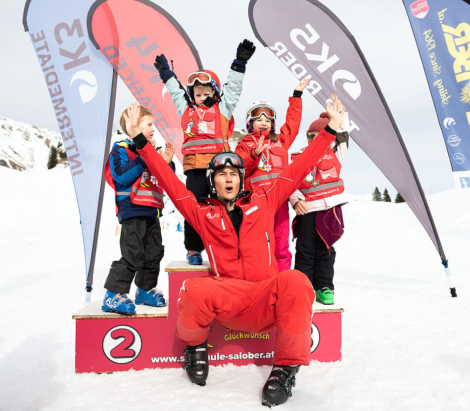 Four cheering children are standing on the podium after the ski race. A ski instructor is seated in front of them with arms wide open and also cheering
