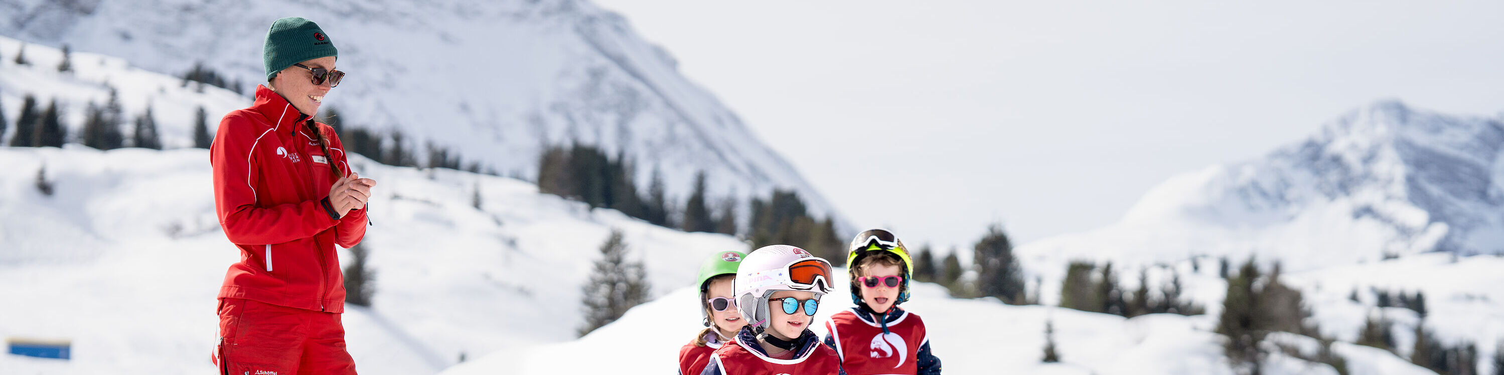 Ein junges Mädchen begibt sich in Startposition beim Kinder-Skirennen, hinter ihm stehen drei andere Kinder und eine Skilehrerin, die applaudiert