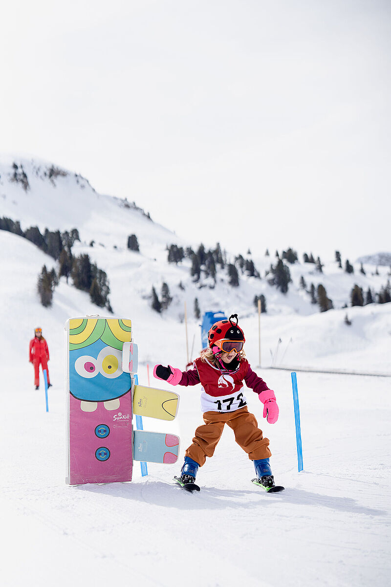Een kind met een lieveheersbeestjeshelm maakt bochten in sneeuwploeg tijdens de skicursus en geeft een high five aan een kleurrijk figuurtje