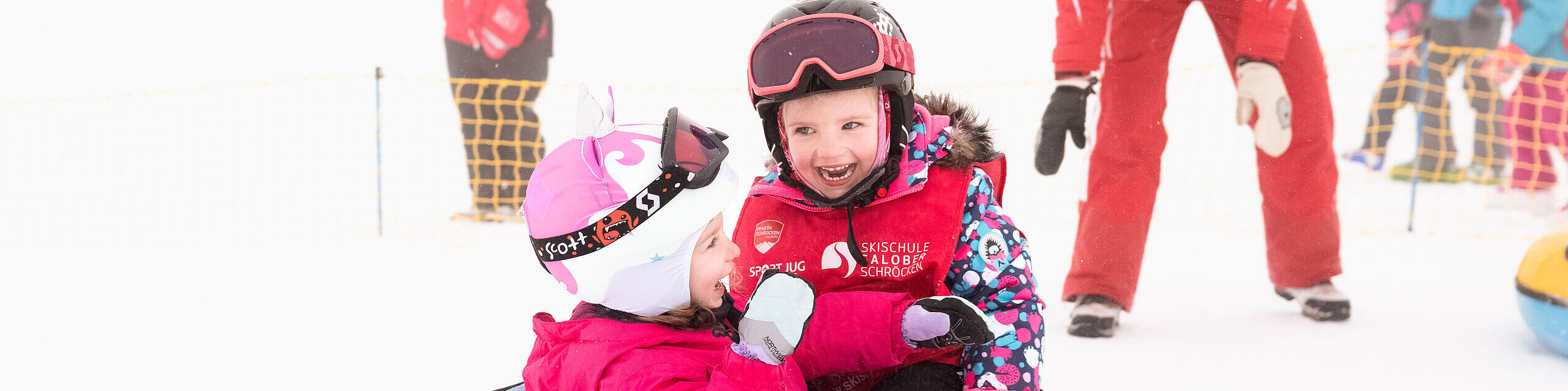 Zwei lachende Kinder sitzen in einem Schnee-Reifen, der von einer Skilehrerin angeschubst wurde