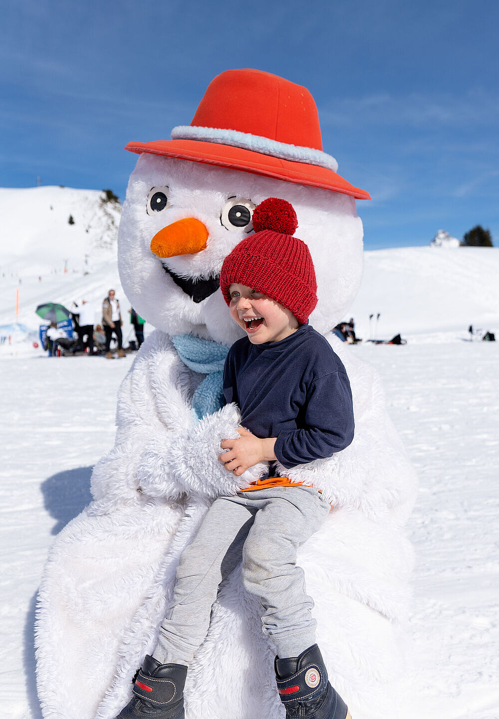 A beaming child with a red cap is sitting on the lap of a snowman mascot