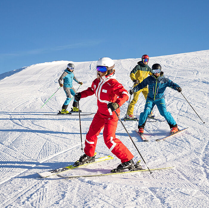 Drei Skifahrer fahren ihrer Skilehrerin auf der Piste im Pflug nach