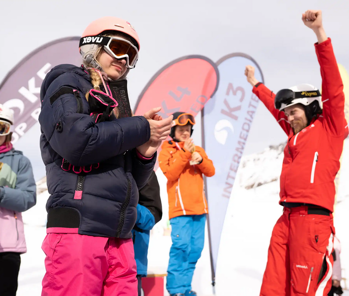 Bij de prijsuitreiking na de skiwedstrijd houden twee jonge deelnemers aan de skicursus een trofee in hun handen en applaudisseren, terwijl de skileraar blij zijn armen in de lucht steekt