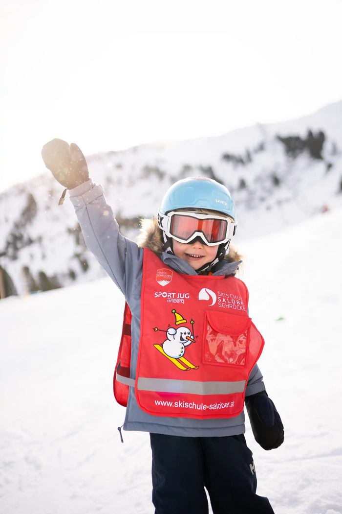 Ein Kind in Ski-Ausrüstung streckt den Arm nach oben und winkt