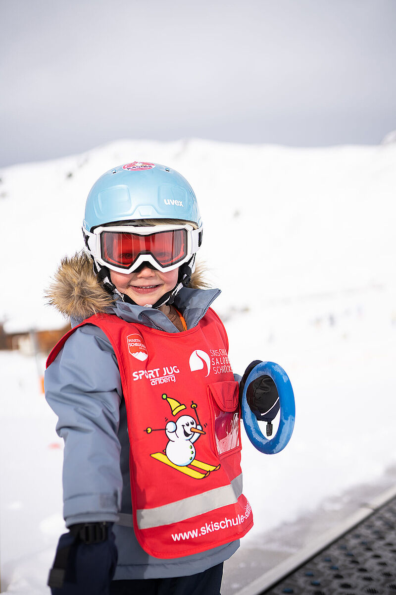 EN Ein fröhliches Kleinkind mit Skibrille, Skihelm und Skikurs-Trikot steht auf dem Zauberteppich