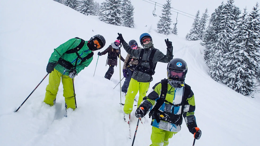 Fünf Jugendliche in Skianzügen, mit Skihelmen und Skibrillen, stehen im Tiefschnee und posen in die Kamera