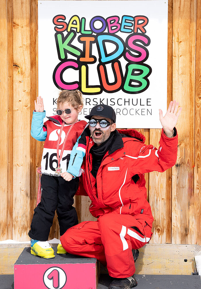 Ein Kind steht auf dem Podium nach dem Skirennen und winkt gemeinsam mit dem Skilehrer in die Kamera