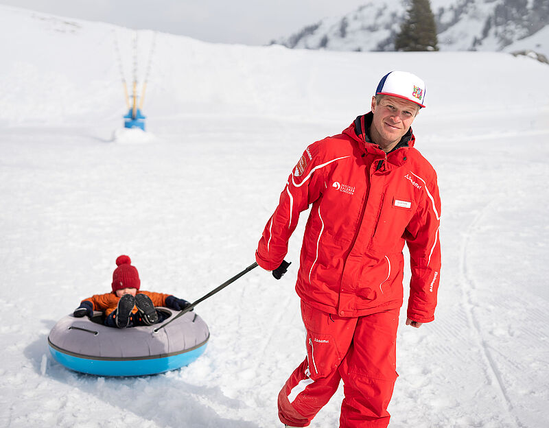Directeur van de skischool Daniel trekt een peuter met een rode muts in een rubberen band op de sneeuw achter zich aan