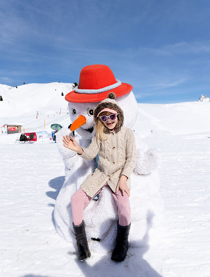 Een kind met een grappige, kleurrijke zonnebril zit op de schoot van sneeuwpop Pauli, de mascotte van de skischool, en zwaait
