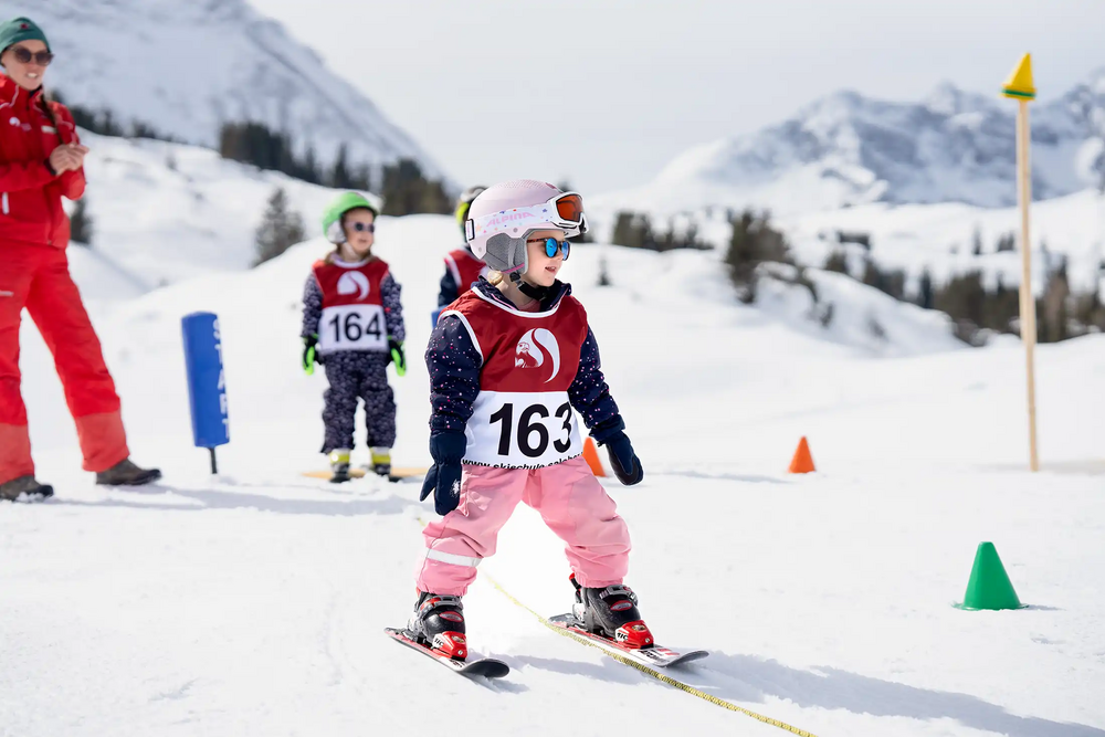 A young girl is getting ready at the start line of a children’s ski race with three other kids and a cheering ski instructor behind her